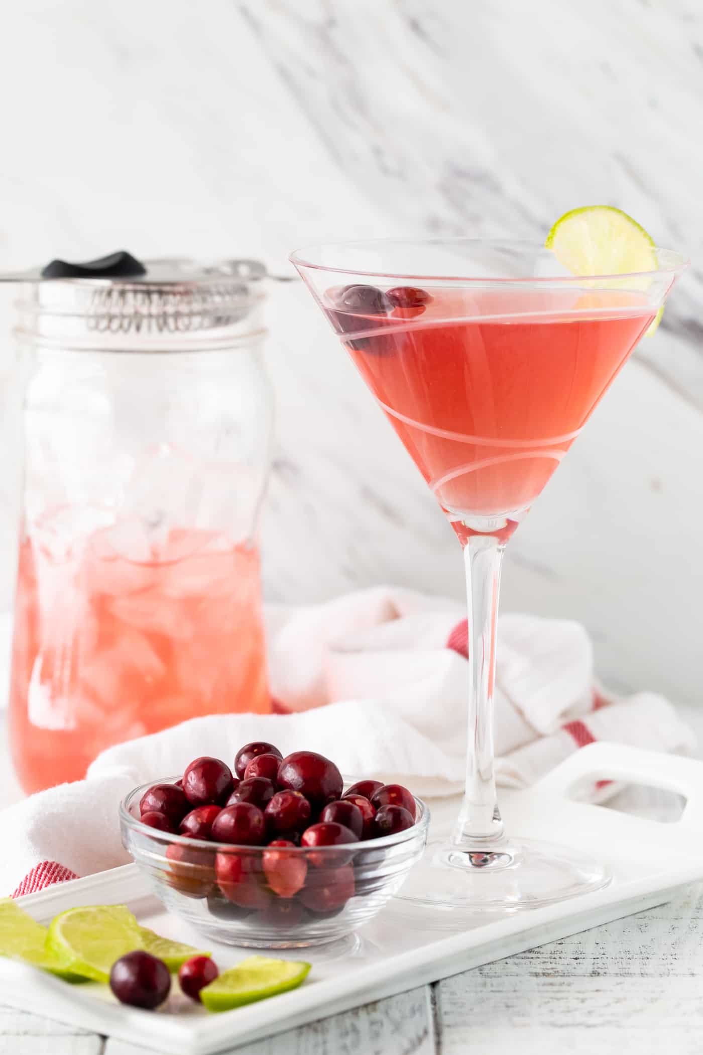 Martini on a white background with a shaker in the background with fresh cranberries and lime wedges.