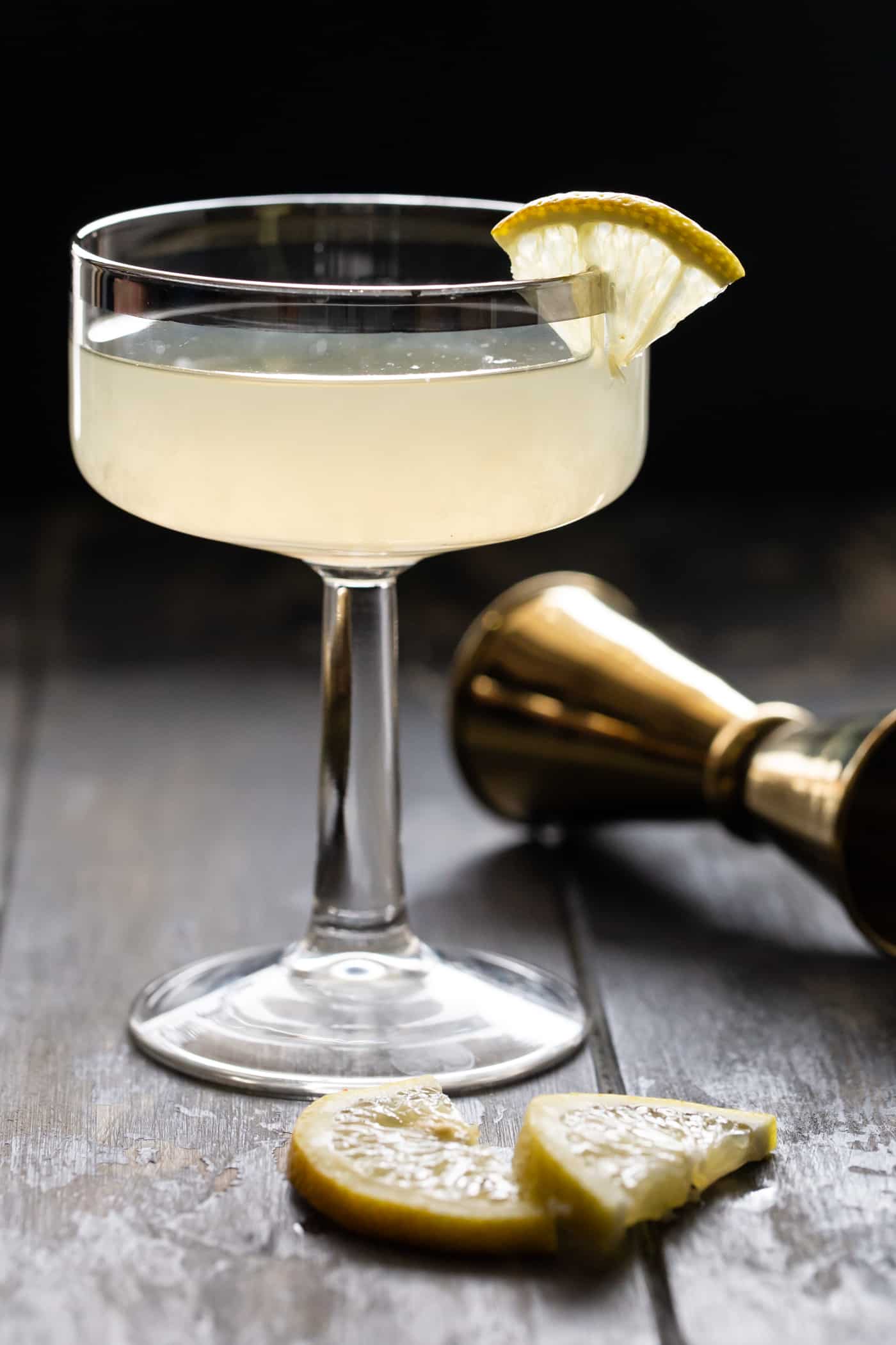 Close up of a  glass filled with Ginger pear Martini recipe and garnished with a small lemon slice. Ona dark background with a gold jigger.
